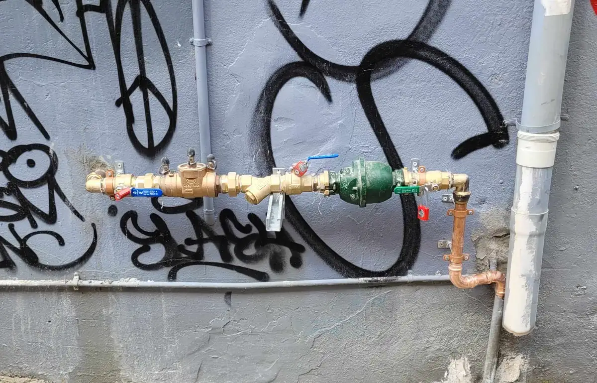 A brass backflow device and fittings mounted along a painted laneway wall covered in graffiti, beside a downpipe