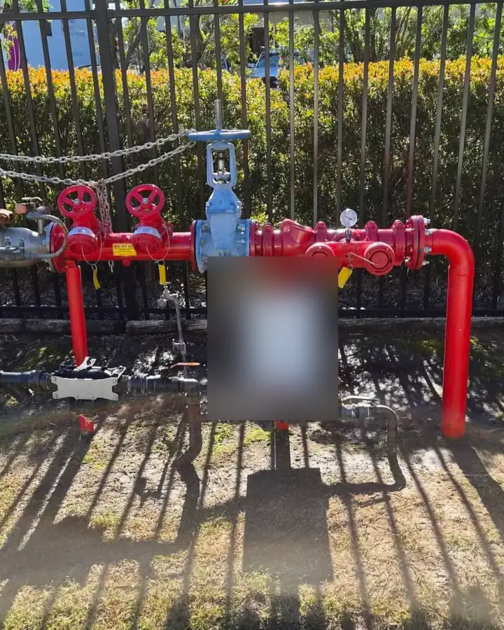 A red booster valve set behind a steel fence, with twin inlets, a blue gate valve and a pressure gauge; the building's board is blurred