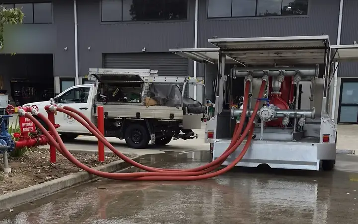 A booster flow test under way: a pump trailer with its side doors open, three red hoses running from its manifold to a fire hydrant booster, a ute parked behind