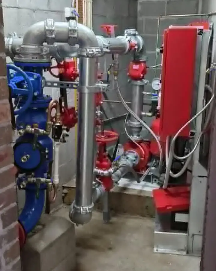 A fire pump room: a red electric pump set with its red controller on the wall, galvanised pipework and a blue valve set beside it