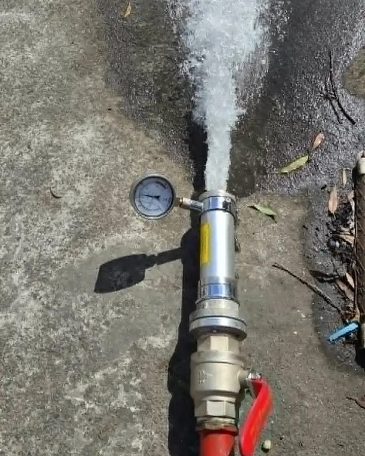 Flow test gear on the ground: water jets out of a steel flow tube with a pressure gauge on its side, fed through a brass valve with a red handle