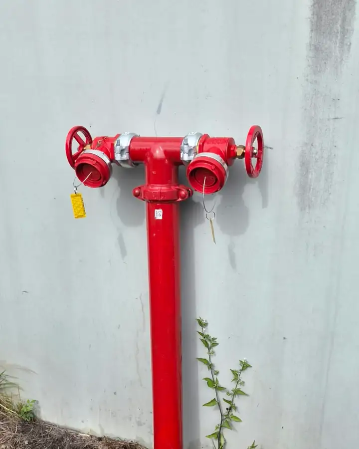 A twin-outlet hydrant against a grey wall, a yellow tag on one outlet