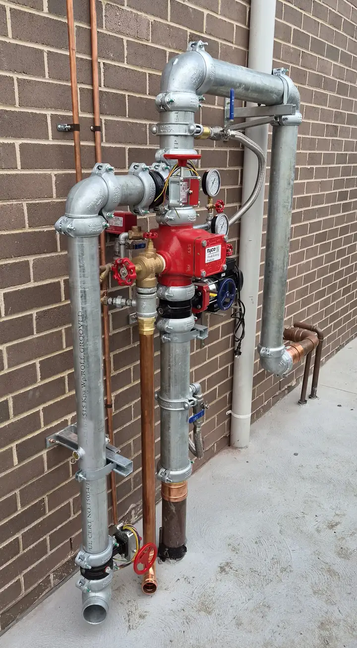 A sprinkler valve set we installed on an outside brick wall: galvanised pipework, the red valve body with its pressure gauges, and red and blue valve handles